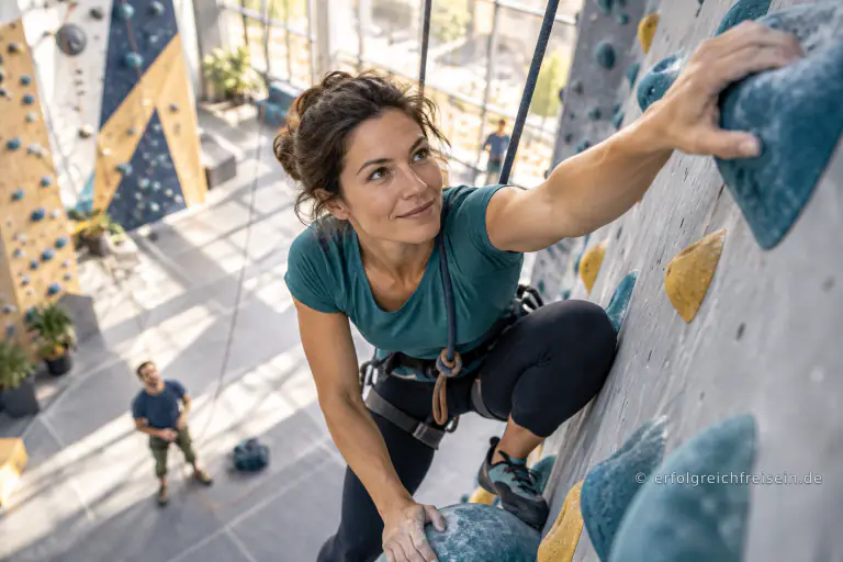 Eine gesicherte Kletterin greift in einer sonnigen Kletterhalle nach dem nächsten Griff.
